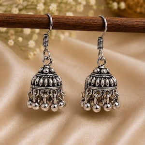 Può includere: Un paio di orecchini jhumka color argento con un design a forma di campana, caratterizzati da intricati motivi in rilievo e piccole perline sferiche pendenti lungo il bordo inferiore. Gli orecchini sono fissati a semplici ganci in metallo.