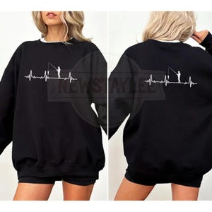 Puede incluir: Sudadera negra con un gráfico blanco de un pescador y una línea de latidos del corazón. El texto "NEWSTAYLEE" es visible en la parte delantera. La sudadera tiene un cuello blanco.