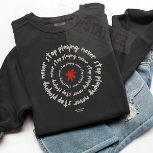 Puede incluir: Sudadera negra con el texto "Never Stop Playing" en un diseño circular. El diseño incluye una flor roja en el centro. La sudadera está doblada sobre unos vaqueros azules.