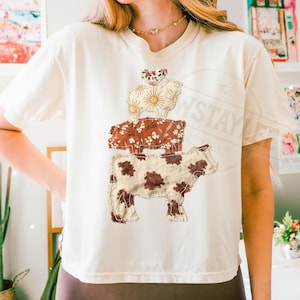 May include: Cream-colored t-shirt featuring a stacked cow design. The design includes three cow shapes, each with a different floral pattern in shades of brown, cream, and red. The text "The Stay" is visible on the right side of the shirt.