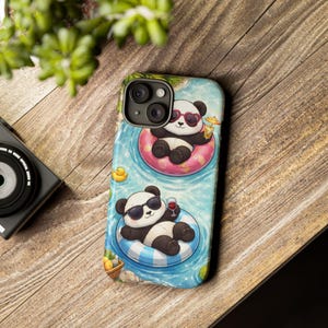 Puede incluir: Una funda de teléfono con una ilustración caprichosa de dos pandas relajándose en una piscina. Un panda lleva gafas de sol en forma de corazón y flota en una boya rosa, mientras que el otro lleva gafas de sol y sostiene una copa de vino.