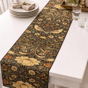 May include: A decorative table runner with a floral and bird pattern. The runner features a dark background with yellow, orange, and green floral designs and two birds. The runner is placed on a white table, adding a touch of elegance to the dining setting.