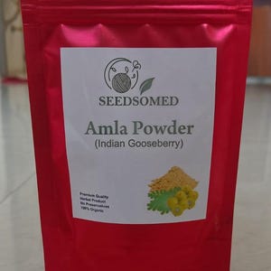 Puede incluir: Una bolsa roja de Amla Powder (grosella espinosa india) de SEEDSOMED. La etiqueta blanca incluye texto y una ilustración del producto. El texto dice "Premium Quality Herbal Product, No Preservatives, 100% Organic."