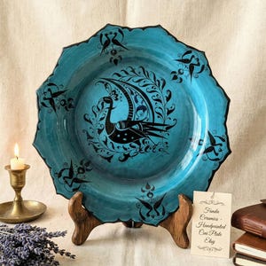 May include: A hand-painted, teal ceramic plate with a black peacock design. The plate has a scalloped edge and is displayed on a wooden stand. A small tag reads "Funda Ceramics - Handpainted Cini Plate Etsy."