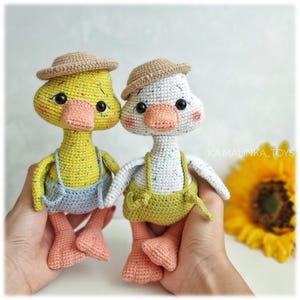 May include: Two handmade crochet duck toys. One is yellow, the other is white. Both ducks wear small brown hats and dungarees. The yellow duck has blue dungarees, and the white duck has green dungarees. They have orange beaks and feet.