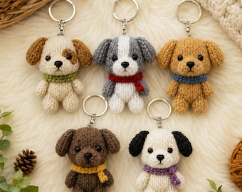 May include: Five small, knitted dog keychains arranged in a group. Each plush puppy features a unique color scheme, including brown, gray, tan, dark brown, and white, with small scarves in green, red, blue, yellow, and purple around their necks.