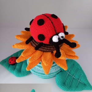 May include: A handmade crochet ladybug perched on a sunflower. The ladybug is red with black spots and has large white eyes. The sunflower has orange petals and a brown center. The base is turquoise and green.