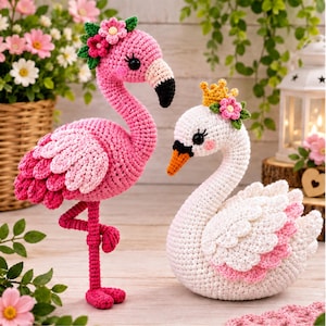 May include: A pink crochet flamingo standing on one leg with a floral headpiece and a white crochet swan wearing a yellow crown and a floral headpiece. Both figures feature detailed stitching, black eyes, and layered wing textures in yarn.