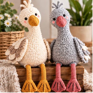 Peut inclure: Deux oiseaux en peluche crochetés à la main assis côte à côte. L'oiseau de gauche est crème avec des accents d'ailes marron et des pattes palmées jaunes. L'oiseau de droite est gris avec un bec rose et des pattes palmées roses. Yeux noirs et joues roses.