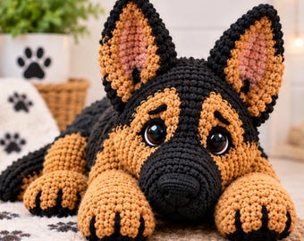 May include: A crocheted German Shepherd puppy toy featuring black and tan yarn, large dark eyes, and upright ears with pink inner details. The plush dog is in a lying position with its front paws extended forward, showcasing detailed black yarn claws.
