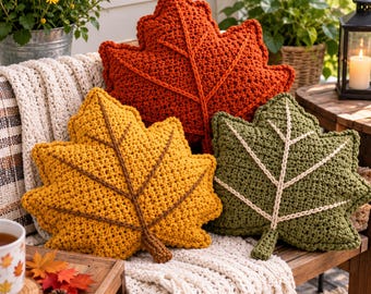 May include: Three leaf-shaped throw pillows made of textured crochet yarn. One pillow is burnt orange with a matching vein, one is mustard yellow with a brown vein, and one is olive green with a cream-colored vein, all featuring a central stem.