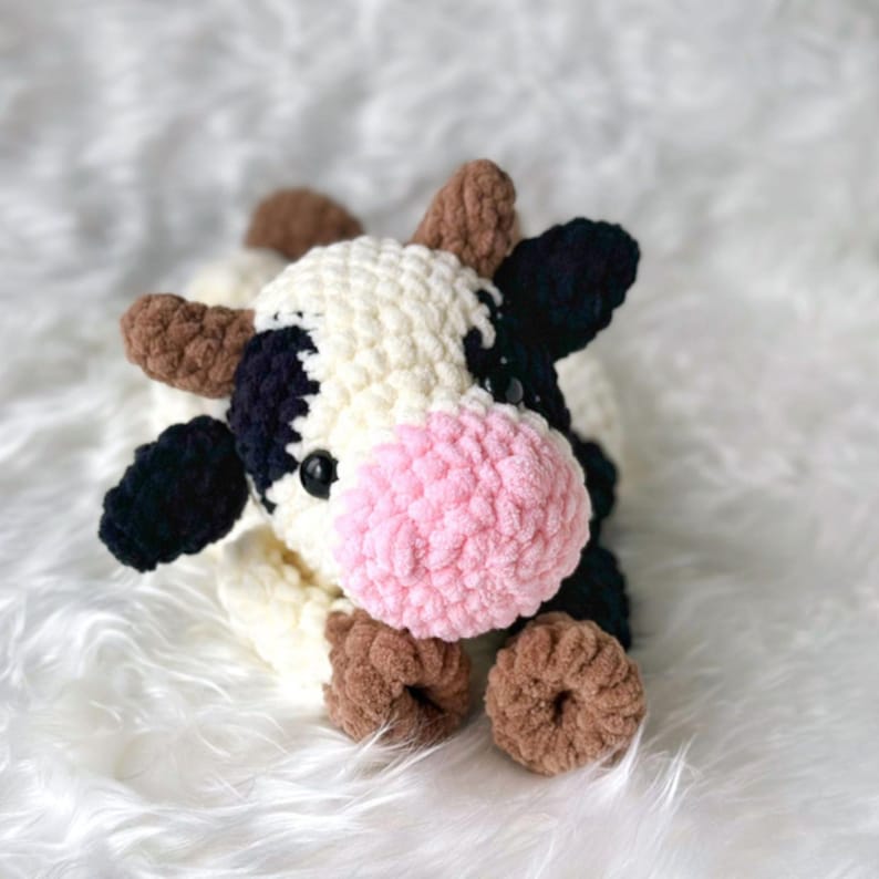 Op de afbeelding: Een handgemaakte gehaakte koeienknuffel met een witte body, zwarte vlekken en bruine hoorns en hoeven. De koe heeft een roze snuit en zwarte ogen. De knuffel is zacht en knuffelig, perfect als kinderspeelgoed of voor de kinderkamer.