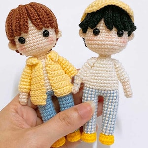 Puede incluir: Dos muñecos de ganchillo hechos a mano. Uno lleva una chaqueta amarilla, vaqueros azules y pelo castaño. El otro lleva una gorra amarilla, una camisa blanca, pantalones a cuadros azules y blancos y pelo negro. Ambos muñecos tienen ojos negros y mejillas rosadas.