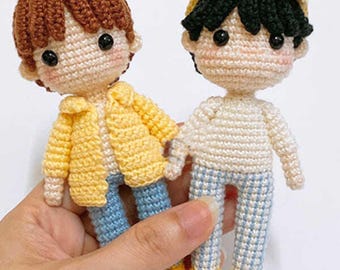 May include: Two handmade crochet dolls. One doll wears a yellow jacket, blue jeans, and brown hair. The other doll wears a yellow cap, white shirt, blue and white checkered pants, and black hair. Both dolls have black eyes and rosy cheeks.