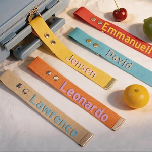 Puede incluir: Una colección de etiquetas de equipaje coloridas con nombres bordados en varios colores. Las etiquetas son amarillas, naranjas, rojas, azules y beige. Una maleta azul está en el fondo. También hay una cereza, una lima y una naranja.