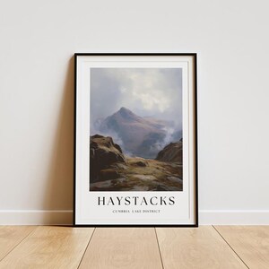 May include: A framed print of Haystacks in the Lake District. The artwork depicts a mountain landscape under a cloudy sky, with rocky terrain in the foreground. The text "HAYSTACKS CUMBRIA LAKE DISTRICT" is printed below the image.