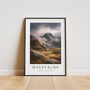 May include: A framed print depicting the Haystacks mountain range in the Lake District, with the text "HAYSTACKS" and "CUMBRIA LAKE DISTRICT". The image shows a misty, overcast sky over a rocky landscape. The print is in a black frame.