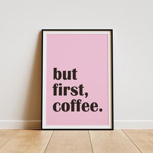 Op de afbeelding: Een ingelijste print met een roze achtergrond en de tekst "but first, coffee" in zwart. De print zit in een zwarte lijst, staand op een houten vloer tegen een witte muur. Eenvoudig, modern ontwerp.