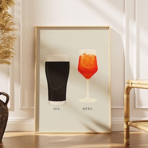 Op de afbeelding: Een rechthoekige wandkunstprint op een lichtbeige achtergrond met een donker stoutbier in een pintglas met het label "HIS." en een feloranje spritz-cocktail met een schijfje sinaasappel in een glas op voet met het label "HERS.".