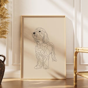 Pode incluir: Uma impressão de arte de parede retangular emoldurada apresentando um desenho de linha contínua preta de um cão em um fundo bege sólido. A obra está em uma moldura de madeira clara, exibindo um estilo artístico canino minimalista.