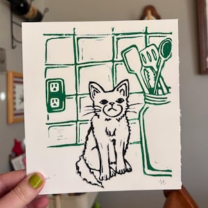 Puede incluir: Un grabado en linóleo que representa un gato frente a una escena de cocina. La impresión es en blanco y negro, con un gato, utensilios de cocina y enchufes. El gato tiene una expresión de mal humor. La impresión está sobre papel blanco.