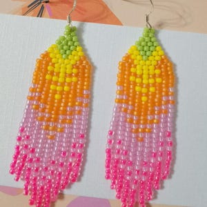 Könnte beinhalten: Ein Paar Perlenohrringe mit Farbverlauf. Die Ohrringe gehen von Grün oben über Gelb, Orange, Rosa bis hin zu Pink unten. Die Ohrringe haben ein fransenförmiges Aussehen.