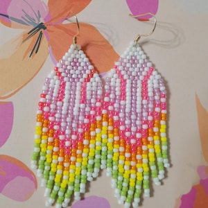 Könnte beinhalten: Bunte Perlenohrringe mit geometrischem Muster. Die Ohrringe weisen einen Farbverlauf auf, darunter Rosa, Weiß, Orange, Gelb und Grün. Die Ohrringe haben goldfarbene Haken.