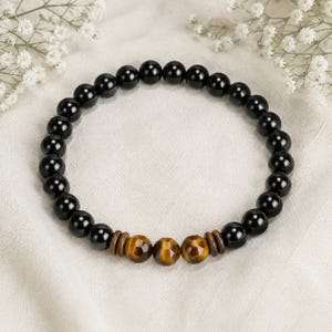 Puede incluir: Una pulsera de cuentas circular con cuentas esféricas negras, lisas y pulidas. El diseño incluye tres cuentas facetadas de ojo de tigre marrón dorado en el centro, flanqueadas por dos pequeños espaciadores cilíndricos de madera marrón oscuro.