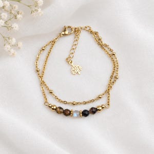 Könnte beinhalten: Ein doppelsträngiges Armband aus goldfarbener Kette mit einer Reihe kleiner facettierter brauner, schwarzer und schillernder Perlen in der Mitte. Das Armband hat einen Karabinerverschluss und eine Verlängerungskette mit einem kleinen baumförmigen Anhänger.