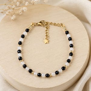 Peut inclure: Un bracelet délicat composé de perles à facettes noires et blanches translucides alternées, agrémenté de petites sphères en métal doré. Le bijou comprend une chaînette d'extension dorée terminée par une petite breloque en forme d'étoile.