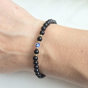 Puede incluir: Una pulsera de cuentas con cuentas negras redondas y lisas dispuestas en un bucle continuo. El diseño incluye un pequeño dije circular de ojo turco azul y blanco, colocado entre dos diminutas cuentas espaciadoras metálicas de tono dorado.