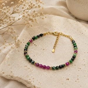 Peut inclure: Un bracelet de perles composé de pierres facettées vertes, noires et magenta, alternées avec de petites perles intercalaires dorées. Le bracelet est doté d'un fermoir mousqueton doré et d'une chaînette d'extension avec une petite breloque.