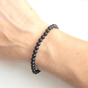 Puede incluir: Una pulsera de una sola hilera compuesta por pequeñas cuentas negras esféricas, lisas y uniformes. Las cuentas presentan una superficie pulida y reflectante que capta la luz. La pulsera está ensartada en un cordón elástico.