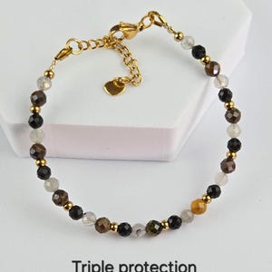 Peut inclure: Un bracelet de perles composé de pierres facettées noires, brunes et gris translucide alternées avec de petites perles métalliques dorées. Le bracelet comprend un fermoir mousqueton doré, une chaînette réglable et un petit cœur. Texte : Triple protection.
