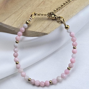 Peut inclure: Un bracelet délicat composé de petites perles roses facettées, entrecoupées de perles dorées. Le bracelet est doté d'une chaîne et d'un fermoir dorés pour un ajustement réglable. Le bijou est présenté sur un tissu blanc et une surface en bois.