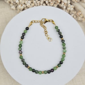Peut inclure: Un bracelet de perles composé de perles rondes vertes et de perles sombres mouchetées, alternées avec de petites sphères en métal doré. Le bracelet comprend un fermoir mousqueton doré, une chaînette de réglage et une breloque en cristal clair.