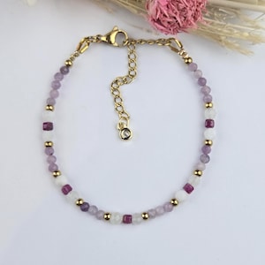 Può includere: Un braccialetto di perline con perline sfaccettate alternate in viola chiaro, bianco e rosa intenso, intervallate da piccole sfere di metallo dorato. Il braccialetto include una chiusura a moschettone dorata e una catenella regolabile con un piccolo ciondolo.