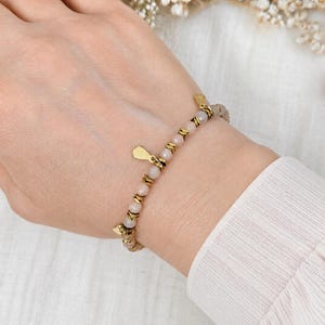 Può includere: Un braccialetto di perline con perline rotonde rosa chiaro alternate a piccoli distanziatori in metallo dorato. Il braccialetto include un piccolo ciondolo piatto a forma di goccia color oro appeso al filo, fissato con una semplice chiusura.