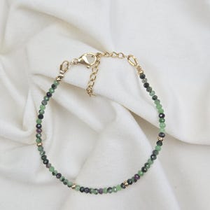 Puede incluir: Una pulsera delicada con pequeñas cuentas de piedras preciosas facetadas en tonos verde, negro y magenta, acentuadas con diminutas cuentas de metal dorado. La pulsera incluye un cierre de mosquetón dorado y una cadena ajustable.