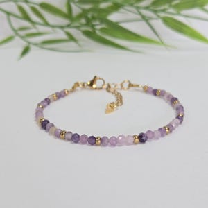Può includere: Bracciale delicato con piccole perline sfaccettate viola e lavanda intervallate da perline color oro. Il bracciale ha una chiusura e una catena regolabile color oro. Lo sfondo è bianco tenue con fogliame verde.