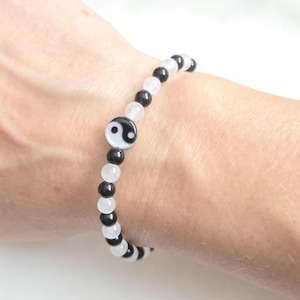 Può includere: Un braccialetto di perline caratterizzato dall'alternanza di perline sferiche nere lisce e bianche traslucide. Il design è centrato su un ciondolo piatto e circolare che mostra un simbolo yin-yang bianco e nero, creando un'estetica equilibrata.