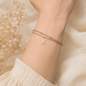 Könnte beinhalten: Ein goldfarbenes Armband mit zwei Strängen, bestehend aus einer feinen Kette mit kleinen integrierten Perlen und einem einzelnen hängenden Anhänger in Form des Großbuchstabens M. Das Schmuckstück hat eine polierte Oberfläche.