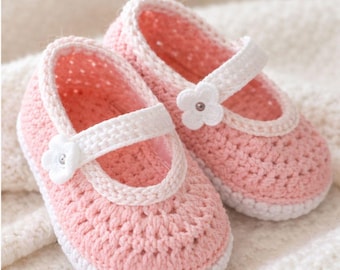 May include: A pair of pink crochet baby shoes with white trim and straps. Each shoe features a white flower accent. The shoes are displayed on a soft, white, textured surface.