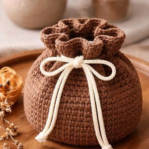 May include: A brown crocheted pouch with a drawstring closure and a cream-colored tie. The pouch is sitting on a wooden tray. The background includes a vase, a candle, and dried flowers.