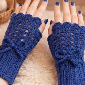 Op de afbeelding: Een paar handgemaakte, blauwe gehaakte vingerloze handschoenen. De handschoenen hebben een geschulpte rand, een openwerkpatroon en een klein strikdetail op de pols. De handschoenen worden getoond op een lichtgekleurd, getextureerd oppervlak.