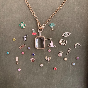 Könnte beinhalten: Eine goldfarbene Halskette mit einem rechteckigen Anhänger und verschiedenen Charms. Zu den Charms gehören ein vierblättriges Kleeblatt, Kirschen, ein Baum, ein Vogel, ein Auge, ein Herz, ein Halbmond, eine Ananas und das Wort "LOVE".