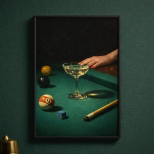 May include: A framed print featuring a still life scene with a champagne coupe on a green pool table. Billiard balls, a cue stick, and a small blue chalk cube are also present. A hand with red nail polish rests on the table.