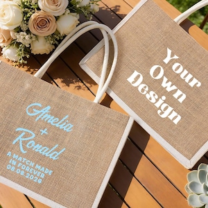 Pu&ograve; includere: Due borse tote in iuta con finiture e manici bianchi. Una borsa ha il testo "Amelia + Ronald" e "A match made in forever 08.08.2026" in azzurro. L'altra borsa ha il testo "Your Own Design" in bianco.