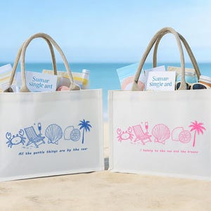 Könnte beinhalten: Zwei weiße Tragetaschen mit Seilgriffen, gefüllt mit Strandutensilien, stehen an einem Sandstrand. Eine Tasche hat blaue Grafiken und den Text "Sumar single art" und "All the gentle things are by the sea." Die andere hat rosa Grafiken und den Text "Saman single art" und "I belong to the sea and the breeze."