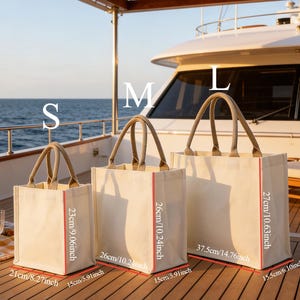 May include: Three canvas tote bags in varying sizes, labeled S, M, and L. The bags have beige fabric, red trim, and jute handles. Measurements are provided in inches: S is 8.27" x 5.91" x 9.06", M is 10.24" x 5.91" x 10.24", and L is 14.76" x 6.10" x 10.63".